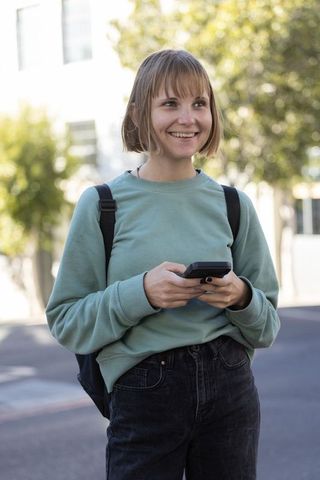 Young woman standing on city street smiling while using smartphone with backpack