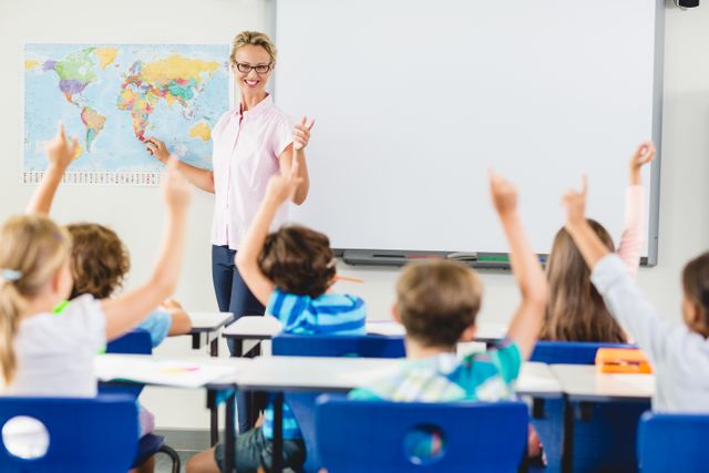 Teacher engaging with students in a classroom setting, pointing to a world map while children raise their hands. Ideal for educational content, school brochures, teaching resources, and articles on classroom interaction and learning environments.
