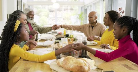 Multi-generation african american family holding hands passing bread sunlit dining table