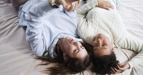 Diverse female friends lying together on bed holding smartphone, sharing quiet moment