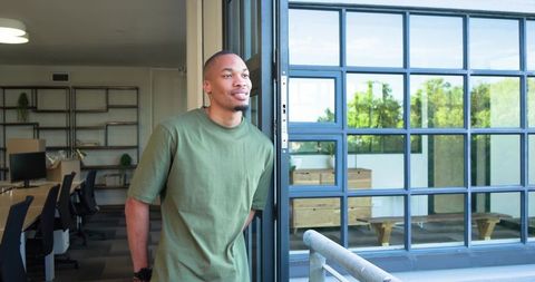 African american man leaning at office balcony door wearing green tee enjoying sunlight