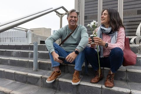 Middle-aged couple sitting on city steps laughing while sharing potted flower and smartphone
