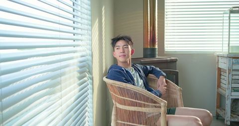 Sunlit asian man relaxing in wicker chair beside window blinds in rustic minimal interior