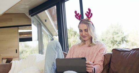 Woman relaxing on sofa wearing reindeer headband and using tablet for holiday browsing