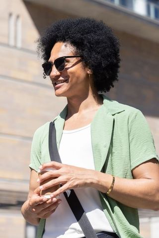 Smiling man in sunglasses green shirt holding coffee with crossbody strap urban sunlight