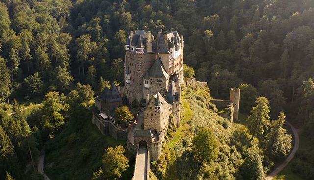 Perching medieval hilltop castle with long stone bridge and ruined tower in dense forest