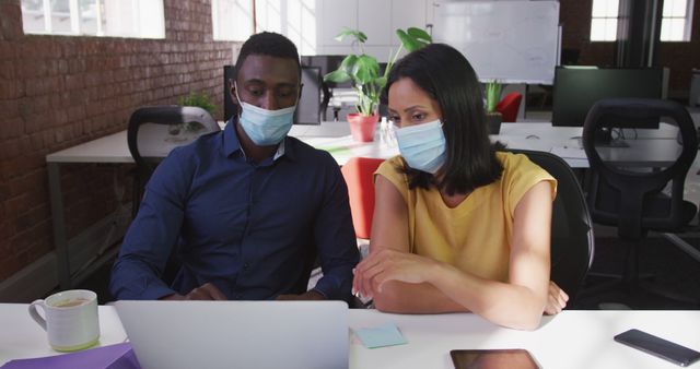 Colleagues collaborating on a project while wearing face masks, demonstrating modern office health protocols. Ideal for depicting safe work environments during health alerts, innovation in teamwork, business cooperation, and contemporary workplace settings.