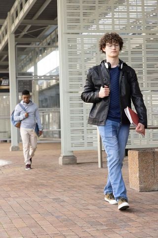 Curly-haired student walking campus carrying red folder and headphones in urban plaza
