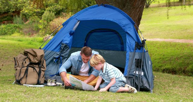 This image depicts a father and son sitting on the grass outside their pitched tent, examining a map together in a park setting. The surrounding area is bright and green with trees and grass, suggesting a serene and pleasant outdoor environment. This type of image is ideal for use in advertisements for outdoor gear, travel guides, family vacation planning brochures, or content promoting family activities and bonding experiences. Businesses related to camping, travel, or parenting can effectively utilize this photo to engage the audience.