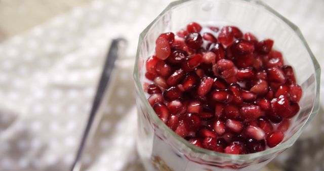 A glass of yogurt topped with fresh pomegranate seeds is placed on a wooden surface, with a spoon resting beside it, with copy space. The vibrant red seeds add a pop of color and hint at a healthy, refreshing snack option.