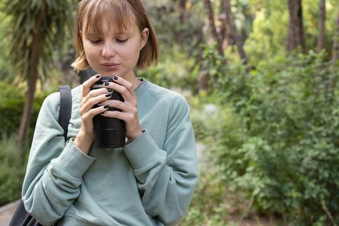 Young woman sipping coffee from travel mug on leafy park trail in mint sweatshirt