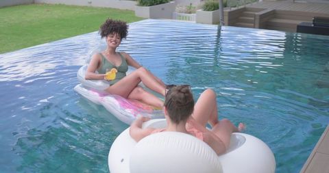 Two women relaxing on colorful inflatable floats in backyard pool with drinks