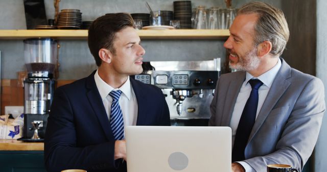 Two businessmen in suits collaborating on a laptop in a coffee shop, discussing business strategies. Ideal for content related to business meetings, teamwork, office discussions, or modern work environments.