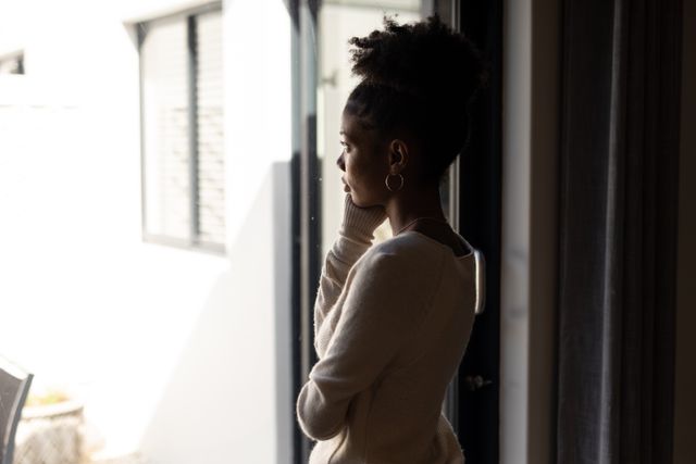 This image captures an African American woman standing by a window, deep in thought. The natural light highlights her contemplative expression, making it suitable for themes of introspection, solitude, and emotional moments. Ideal for use in articles or advertisements related to mental health, lifestyle, personal growth, and home living.