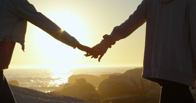 Two people are holding hands against a beautiful sunset backdrop by the sea, with copy space. Their silhouette and the warm glow of the setting sun create a romantic and peaceful atmosphere.