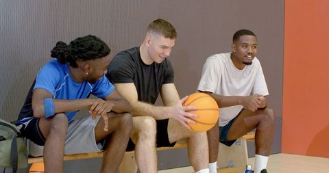 Diverse basketball teammates chatting on bench holding ball after practice, relaxing