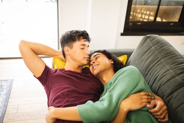 This image depicts a romantic multiracial gay couple cuddling and lying on a sofa in a living room. It can be used for promoting LGBTQ inclusivity, relationship counseling, lifestyle blogs, home decor, and advertisements focusing on love and togetherness.