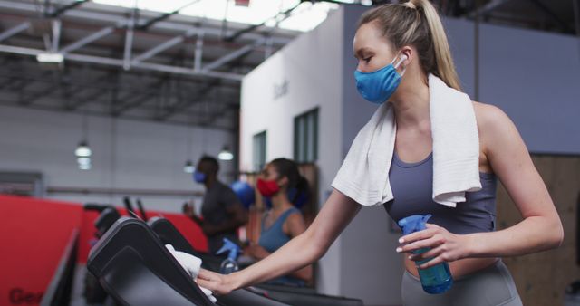 Woman exercising at a gym is cleaning a treadmill while wearing a face mask. The towel and spray bottle emphasize importance of hygiene and health safety in shared spaces. This image can be used for promoting clean and safe workout environments, gym hygiene protocols, or fitness routines during a pandemic.