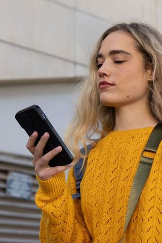 Young woman checking phone in mustard sweater with backpack strap urban street portrait