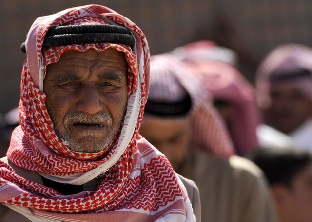 Elderly Middle Eastern man wearing traditional attire stands in an outdoor group setting with others in the background. The scene evokes themes of cultural heritage, community gatherings, and traditional customs. Suitable for use in articles or projects discussing Middle Eastern traditions, elderly lifestyles, cultural diversity, or communal activities.
