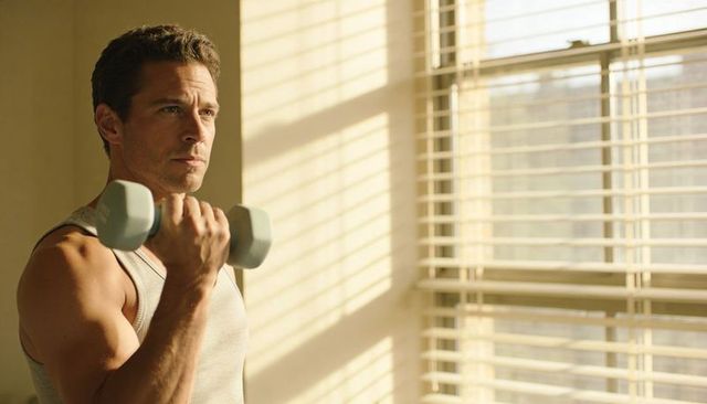 Man lifting dumbbell at home in morning sunlight performing bicep curl by window blinds