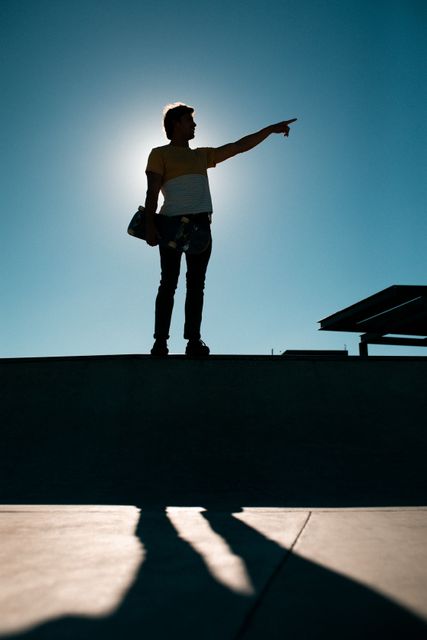 Silhouette of a man holding a skateboard and pointing at something while standing at an urban skatepark during summer. Ideal for use in advertisements, lifestyle blogs, sports magazines, and promotional materials related to skateboarding, outdoor activities, and youth culture.