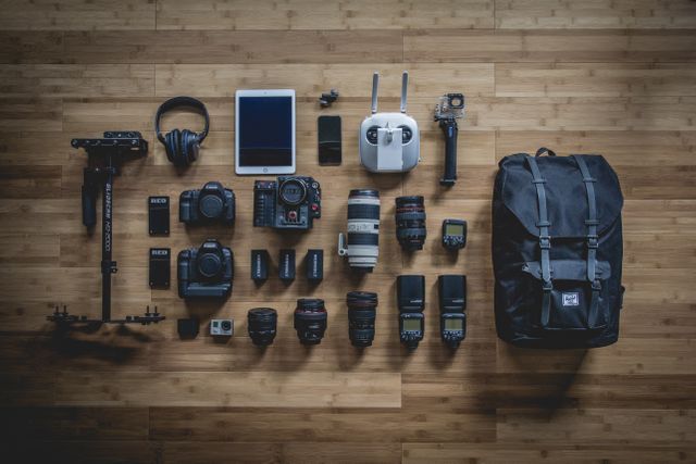 Various photography and videography tools neatly arranged on wooden floor. Includes cameras, lenses, gimbals, headphones, and travel backpack, demonstrating readiness for professional photoshoots and video recording. Perfect visual for photography blogs, travel sites, or equipment reviews.