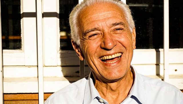 Laughing senior man squinting in bright porch sunlight, warm portrait closeup, joyful expression