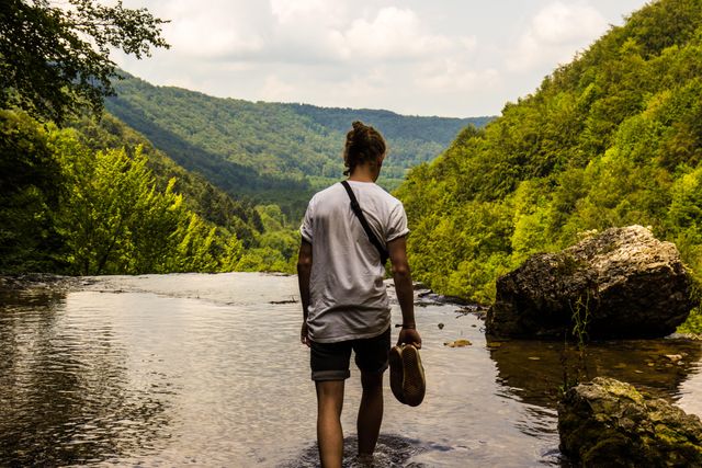 Man walking in shallow river barefoot in mountainous area with lush green hills and forest. Perfect for nature-related articles, travel tips, and adventure blog posts. Great for use in campaigns promoting outdoor activities, hiking spots, and eco-tourism tours.