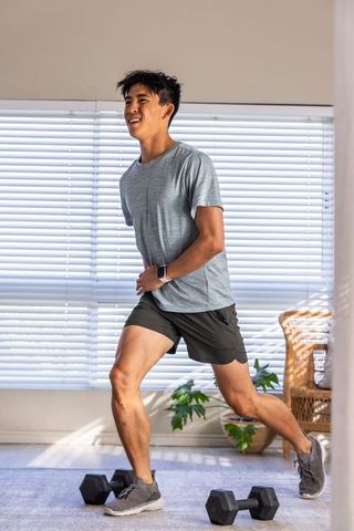 Asian man performing lunge during sunlit home workout wearing smartwatch with dumbbells