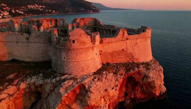 Glowing sunset fortress ruins on rugged cliff with round tower and coastal battlements