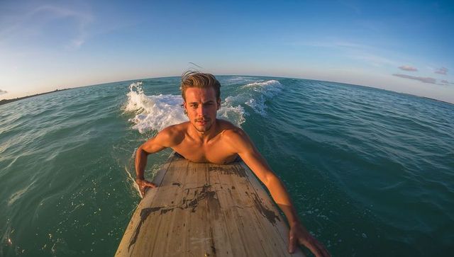 Shirtless surfer paddling on surfboard in turquoise ocean at sunrise, wave breaking, adventure