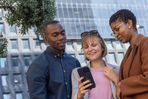 Diverse professionals leaning in, smiling and sharing smartphone outside glass office facade