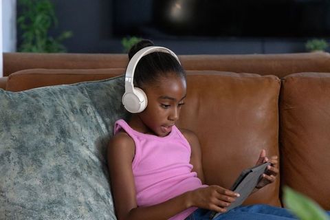 African-american girl wearing white headphones using tablet on brown couch, focused learning