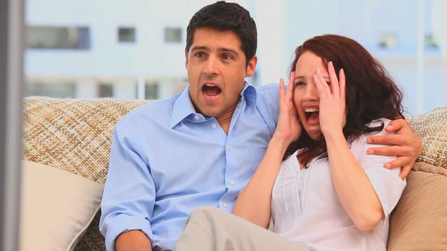 Couple sitting on sofa displaying expressions of fear and surprise while watching a scary movie. Could be used for entertainment advertisements, social media posts about movie nights, or articles discussing film reactions at home. Perfect for illustrating bonding activities or reactions to entertainment.