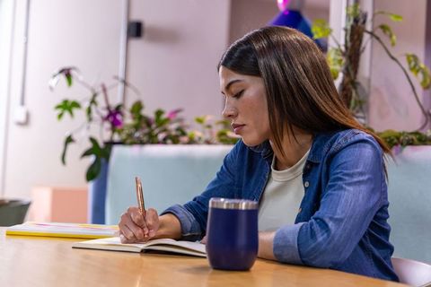 Young woman writing in notebook at coworking lounge with plants, tumbler and pastel decor
