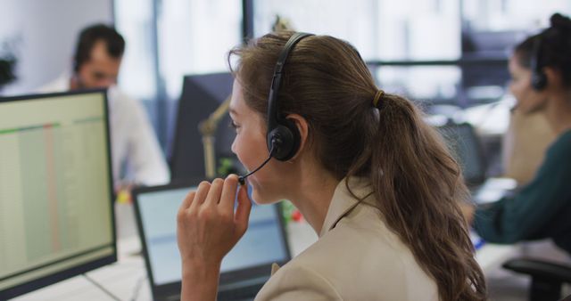 Customer service agent wearing headset working diligently at computer desk in busy professional environment. An image ideal for illustrating concepts of business communication, office teamwork, client support, and professional engagement. Suitable for corporate websites, customer service training materials, and business-related presentations.