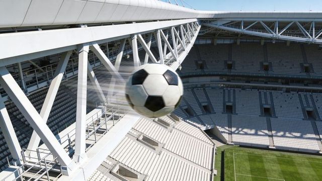 Soaring soccer ball arcing over modern stadium with exposed steel trusses and empty seats