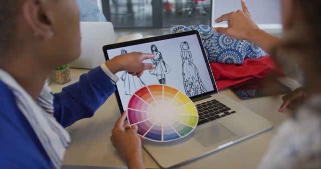 Fashion designers actively collaborating on a sketch on a laptop screen. One designer is holding a color palette while pointing at the sketches on the screen. This visual is perfect for illustrating creative teamwork, the design process, and design collaboration in the fashion industry. Suitable for use in articles, blogs, or websites discussing fashion design, creative processes, and professional collaboration.