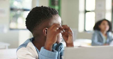 African american teen rubbing eyes at classroom desk with laptop and blurred peer behind