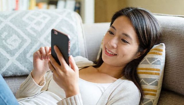 Smiling woman reclining on sofa using smartphone, cozy living room, casual cardigan comfort