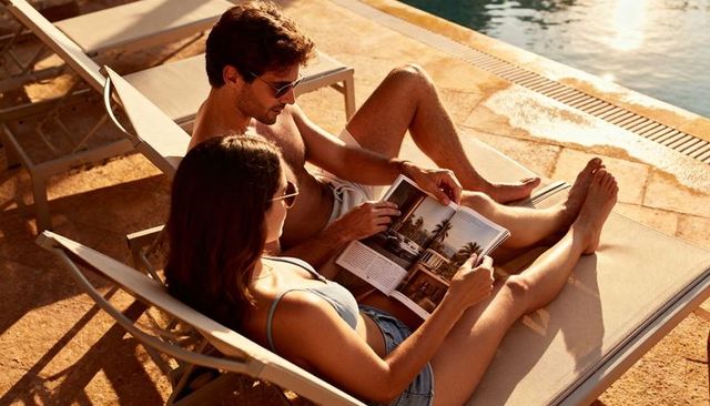 Sunlit couple reading magazine on poolside loungers, relaxing vacation lifestyle scene