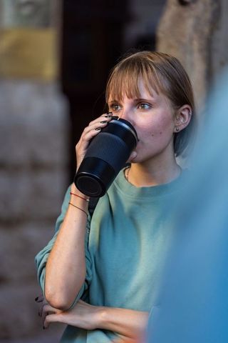 Young woman drinking from black reusable travel mug while wearing teal sweatshirt outdoors