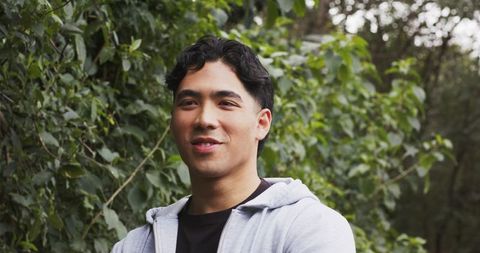 Young Asian man standing outdoors smiling in gray hoodie against green foliage