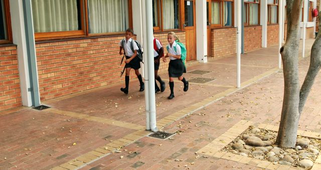 Three young schoolchildren in uniforms with backpacks walking together outside a brick classroom building. The scene represents education, friendship, and the school journey. Ideal for use in educational materials, advertisements for school supplies, and articles about student life and the education system.