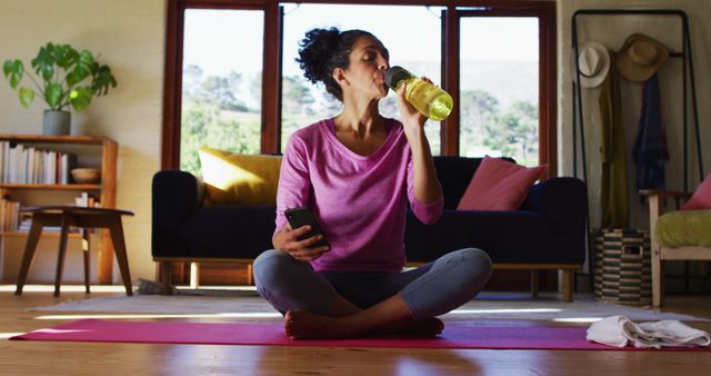 This stock photo shows a woman sitting on a yoga mat in a modern living room, drinking from a bottle of water while holding a smartphone. She appears to be taking a break during her home workout session. She is wearing comfortable casual clothing. This image is ideal for use in articles, blogs, and advertisements related to home fitness, hydration, self-care, and healthy lifestyles.