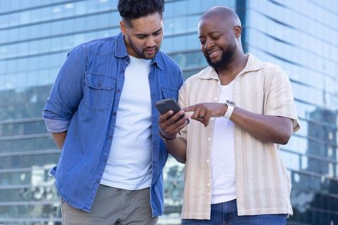 Two men sharing smartphone and smiling outside modern glass office building