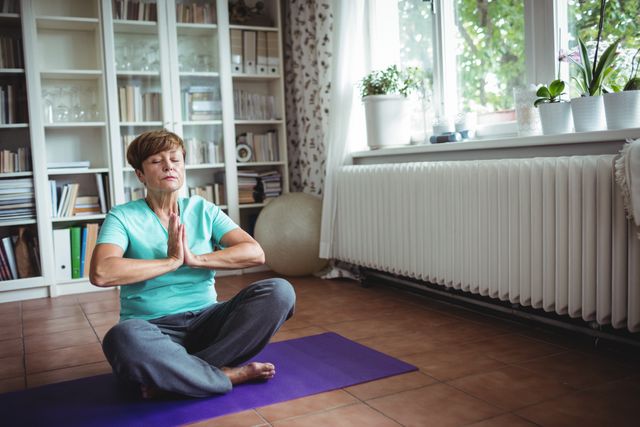 Senior woman sitting on yoga mat meditating in prayer position at home. Ideal for content related to senior wellness, mindfulness practices, home workouts, mental health, and self-care routines. Can be used in articles, blogs, and advertisements promoting healthy lifestyles for the elderly.