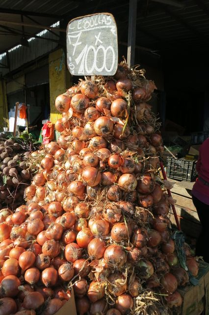 Large pile of fresh onions displayed at outdoor farmer's market with a sign indicating price of 7 for 1000. Ideal for use in content related to agriculture, fresh produce, markets, vegetable stalls, and healthy eating. Can be used in blogs, articles, advertisements, and promotional materials for farmer's markets or local produce shops.