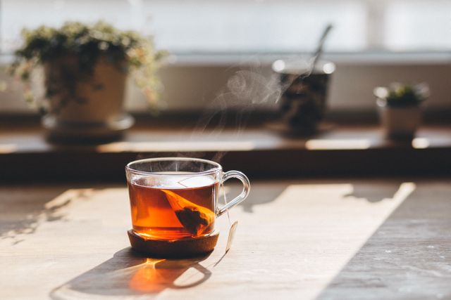 Steaming cup of tea sitting on a wooden table in a sunlit kitchen. Sunlight pours through the window, casting soft shadows, creating a warm and inviting atmosphere. Ideal for themes related to relaxation, home comfort, morning routines, and peace.